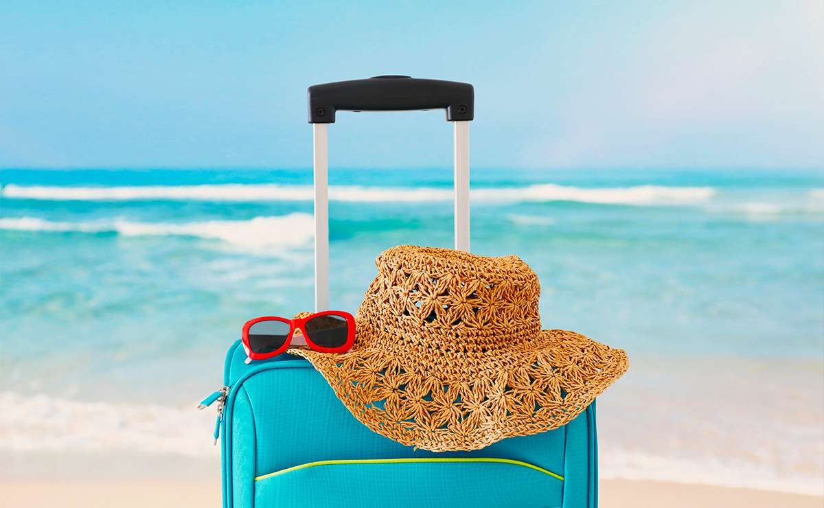 Valigia azzurra su una spiaggia con cappello di paglia e occhiali da sole rossi appoggiati sopra, con il mare sullo sfondo.