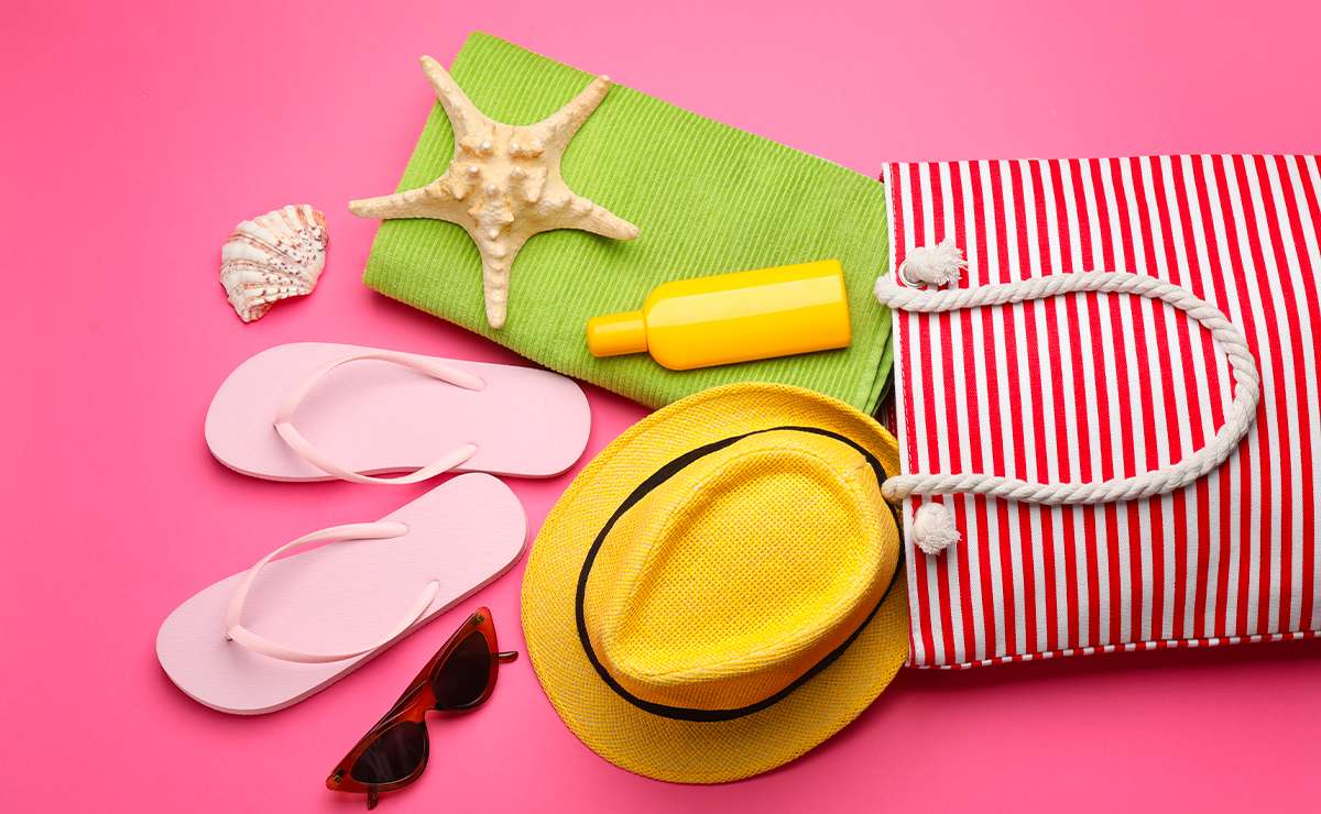 Accessori da spiaggia su sfondo rosa: borsa a righe rosse, infradito rosa, cappello giallo, occhiali da sole, protezione solare, stella marina e asciugamano verde.