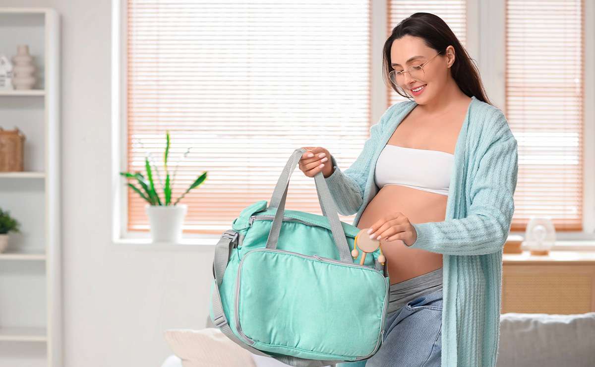 Donna incinta sorride mentre prepara la borsa per il parto in casa, indossando abiti comodi e cardigan azzurro, con una valigia ospedaliera verde acqua in mano.