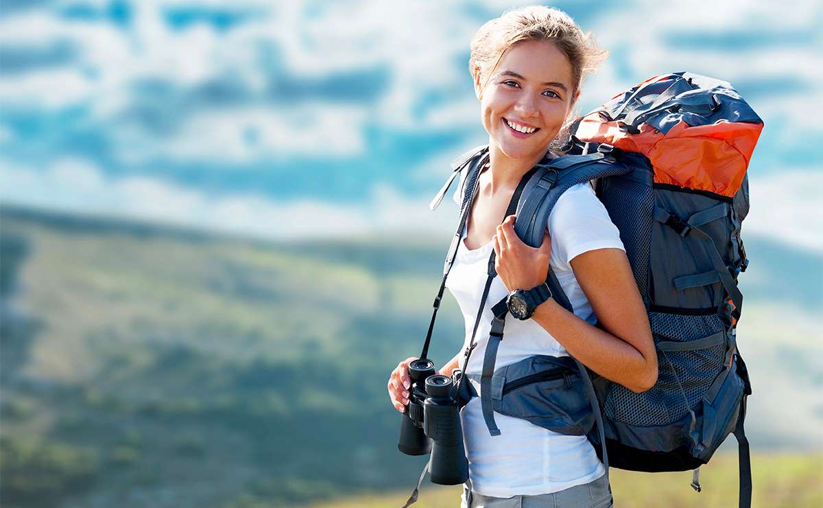 Donna sorridente con zaino da trekking, binocolo e abbigliamento tecnico in montagna durante un’escursione estiva.