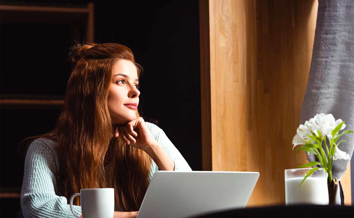Donna con lunghi capelli castani seduta accanto a un laptop, guarda pensierosa fuori dalla finestra mentre sorseggia un caffè.