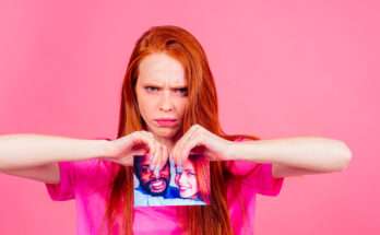 Donna dai capelli rossi con espressione arrabbiata che strappa una foto di coppia davanti a sfondo rosa.