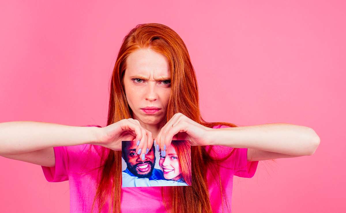 Donna dai capelli rossi con espressione arrabbiata che strappa una foto di coppia davanti a sfondo rosa.
