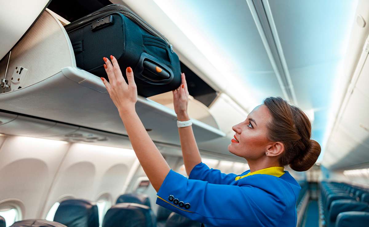 Assistente di volo in uniforme ripone un trolley nero nella cappelliera di un aereo