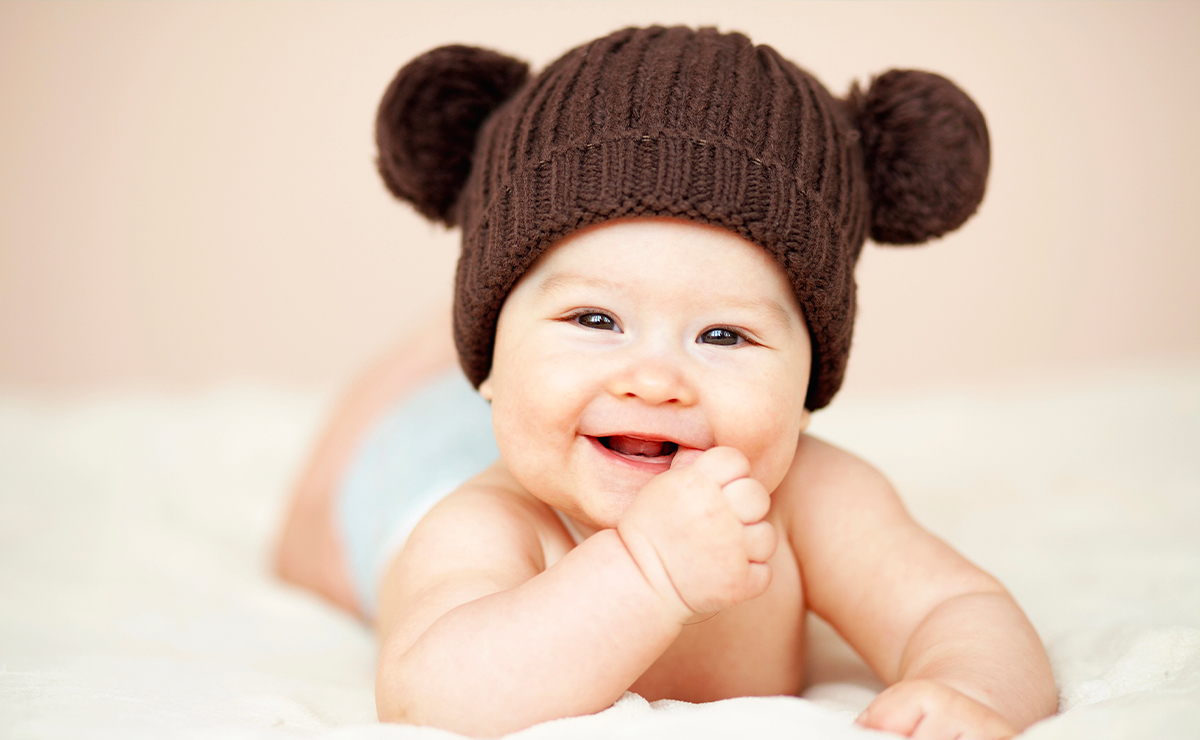 Bambino sorridente con cappellino marrone a forma di orsetto, disteso su un letto chiaro