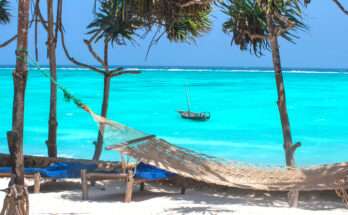 Amaca in spiaggia all’ombra delle palme con vista su mare turchese a Zanzibar, con dhow tradizionale in lontananza e lettini sulla sabbia bianca.