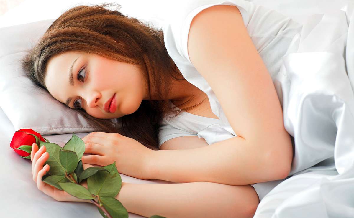 Donna distesa su un letto con espressione triste, vestita di bianco, mentre tiene tra le mani una rosa rossa