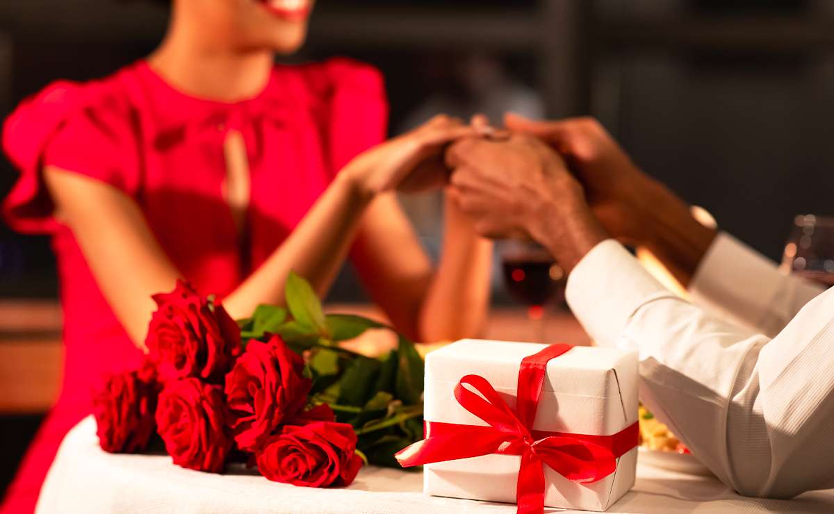 Cena romantica con donna in abito rosso che sorride mentre tiene le mani a un uomo, con rose rosse e un regalo con fiocco in primo piano