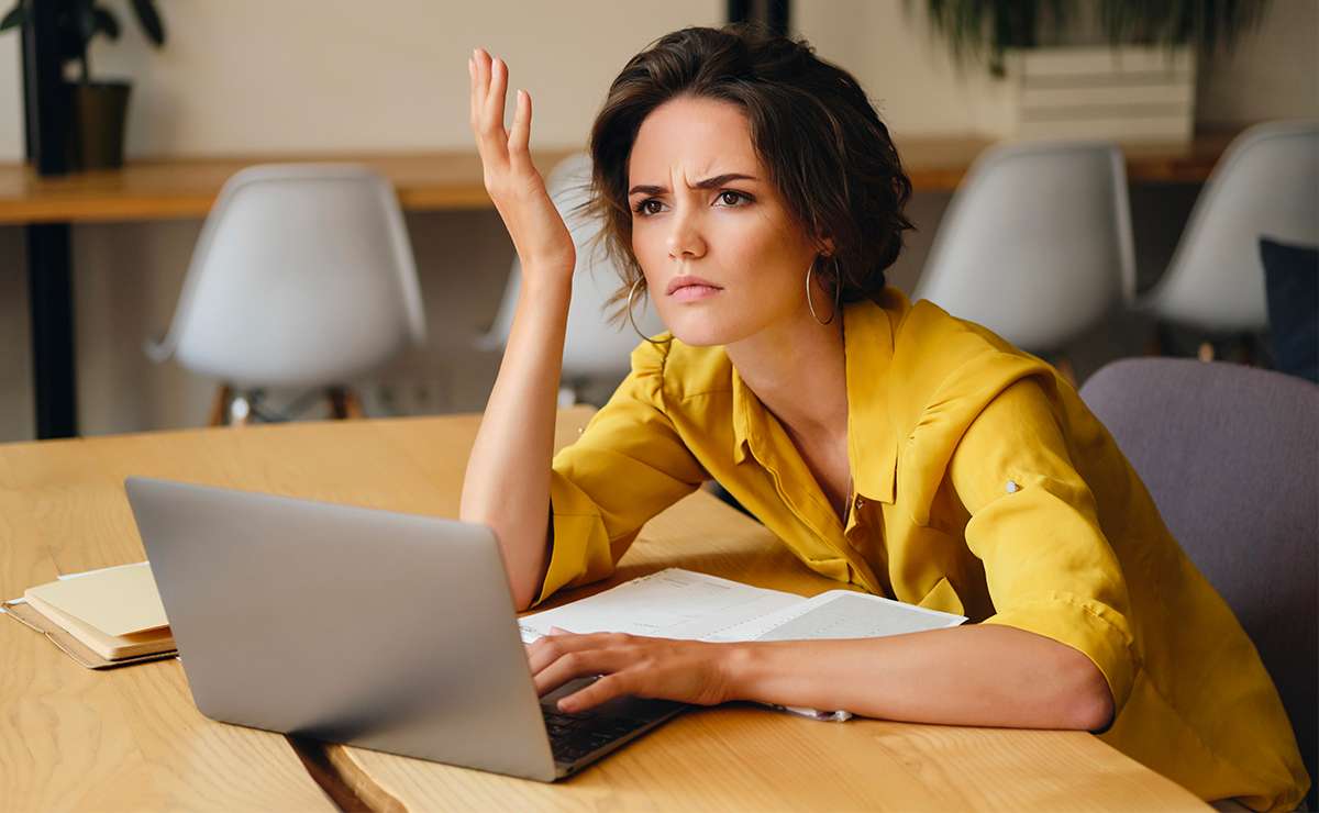 Donna con camicia gialla seduta a una scrivania, davanti a un computer portatile, con espressione perplessa e una mano alzata in segno di frustrazione, mentre guarda lo schermo. Sul tavolo ci sono un’agenda e un blocco appunti. La scena trasmette senso di confusione e disagio in un contesto lavorativo.