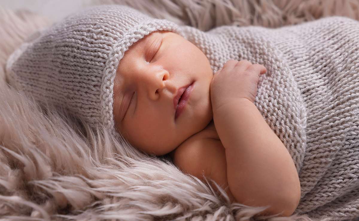 Neonata che dorme avvolta in una copertina lavorata a maglia color panna, sdraiata su un morbido plaid beige, in primo piano.