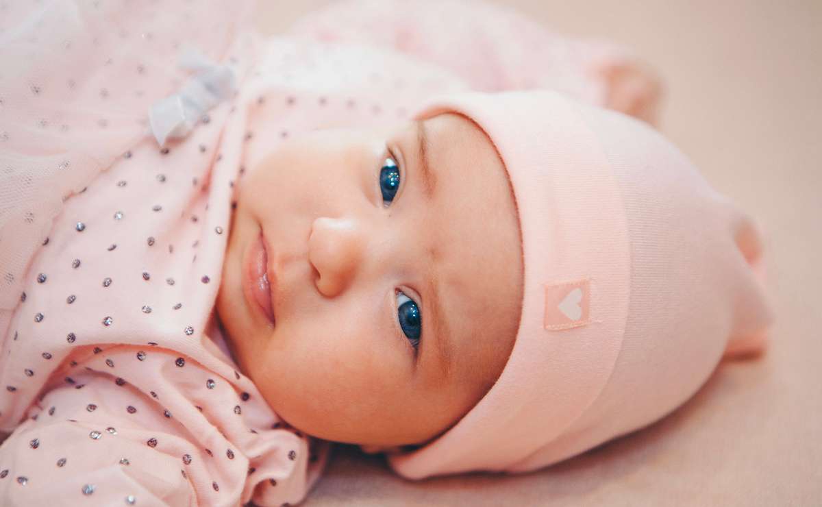 Neonata con occhi azzurri e berretto rosa, vestita con tutina rosa a pois e sdraiata su una coperta chiara, in primo piano.