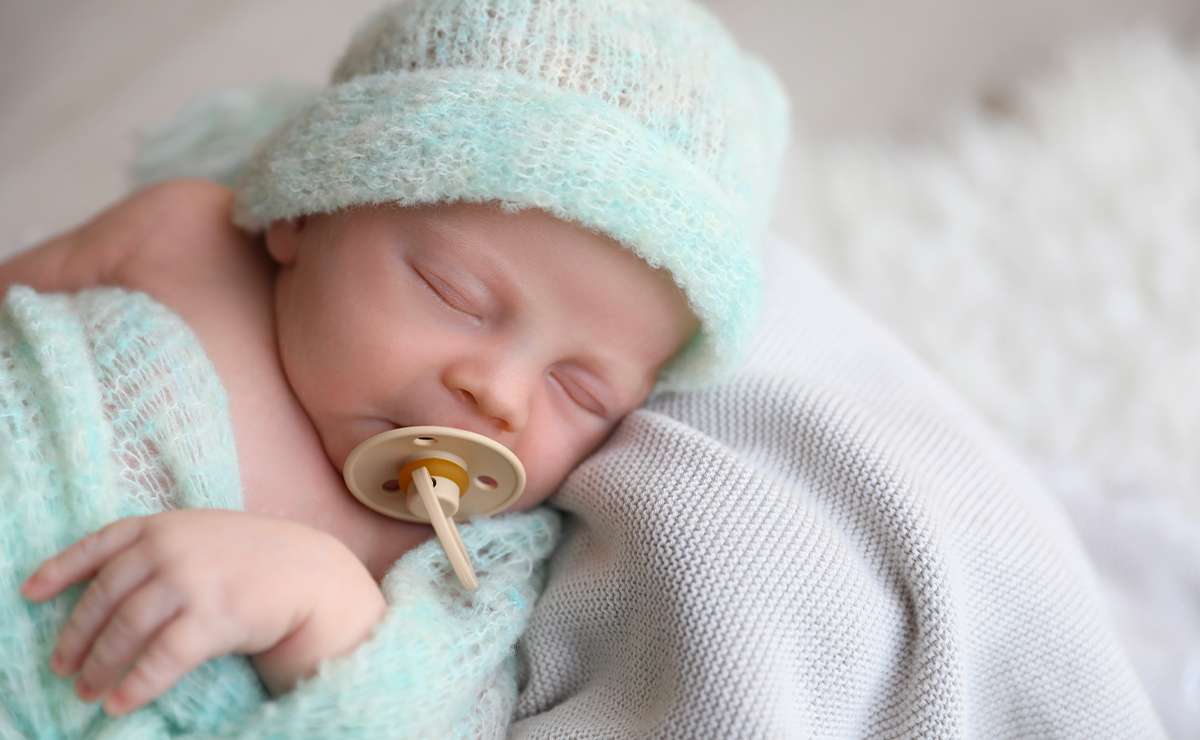 Neonato che dorme con ciuccio, avvolto in una copertina verde acqua e berretto coordinato, sdraiato su una coperta in tessuto chiaro.