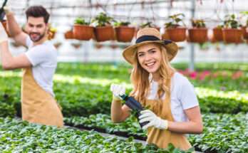 Due giovani sorridenti impegnati in attività di ortoterapia in serra, intenti a curare le piante con guanti e attrezzi da giardinaggio terapeutico.