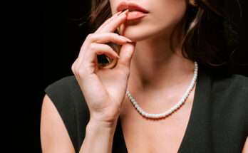 Donna elegante con collana di perle bianche indossa un abito nero, con mano vicino alle labbra in posa riflessiva su sfondo nero scuro.