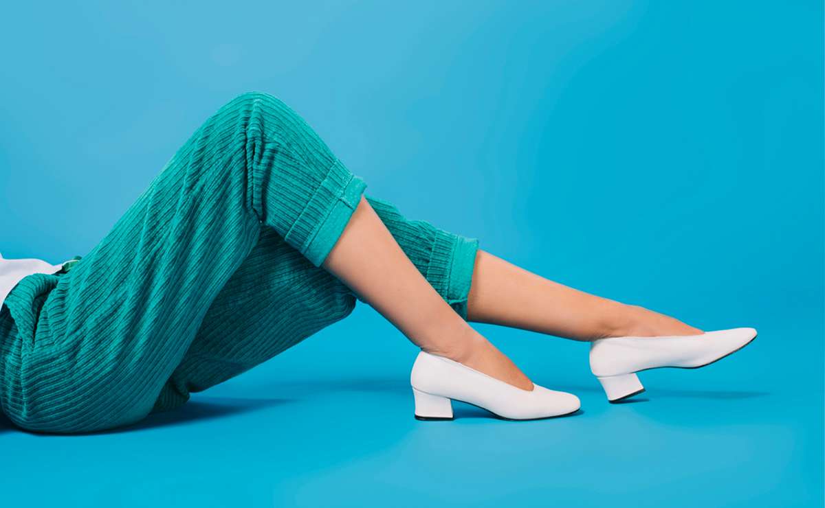 Gambe femminili con pantaloni verde smeraldo e décolleté bianche con tacco basso su sfondo azzurro, concetto di stile elegante e comfort.