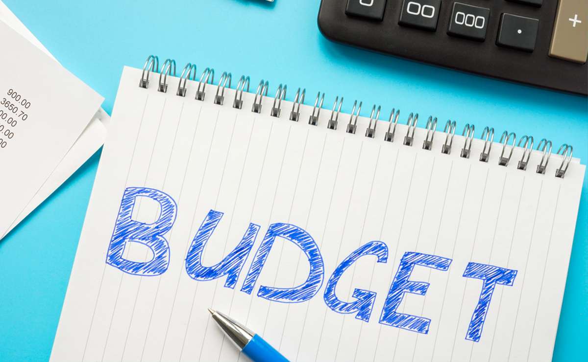 Quaderno con la scritta “BUDGET” in primo piano, accompagnato da una penna, calcolatrice e ricevute su sfondo azzurro, concetto di gestione delle finanze personali.