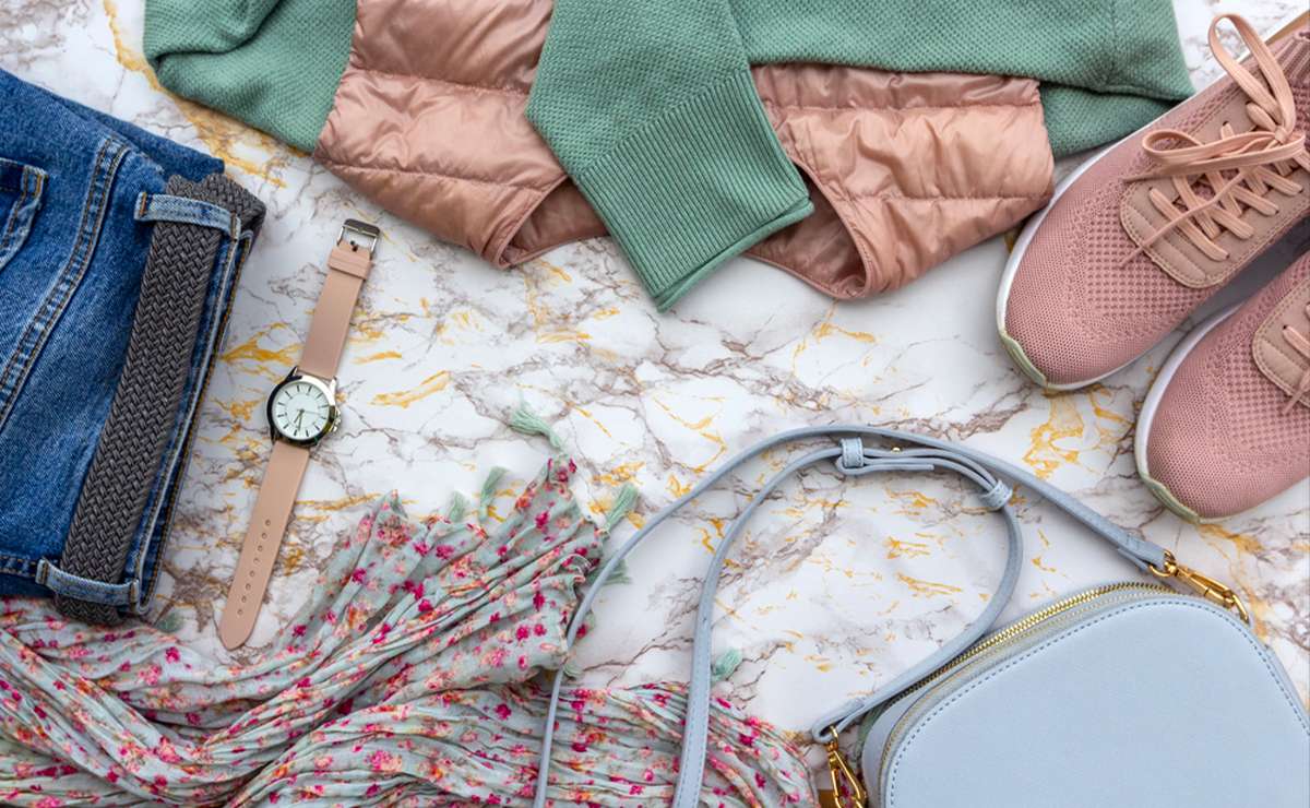 Composizione flat lay di abbigliamento e accessori femminili per la primavera-estate 2025: jeans, maglia verde con dettagli rosa cipria, scarpe sportive rosa, orologio beige, borsa a tracolla azzurra e foulard floreale su sfondo marmorizzato.