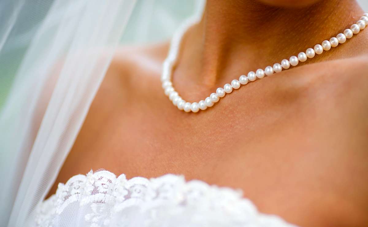 Dettaglio del collo di una sposa con una collana di perle bianche e abito da matrimonio in pizzo, su sfondo sfocato.