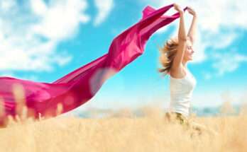 Donna felice in un campo dorato con foulard fucsia al vento sotto il cielo estivo: simbolo di libertà, benessere e cura del corpo in estate.