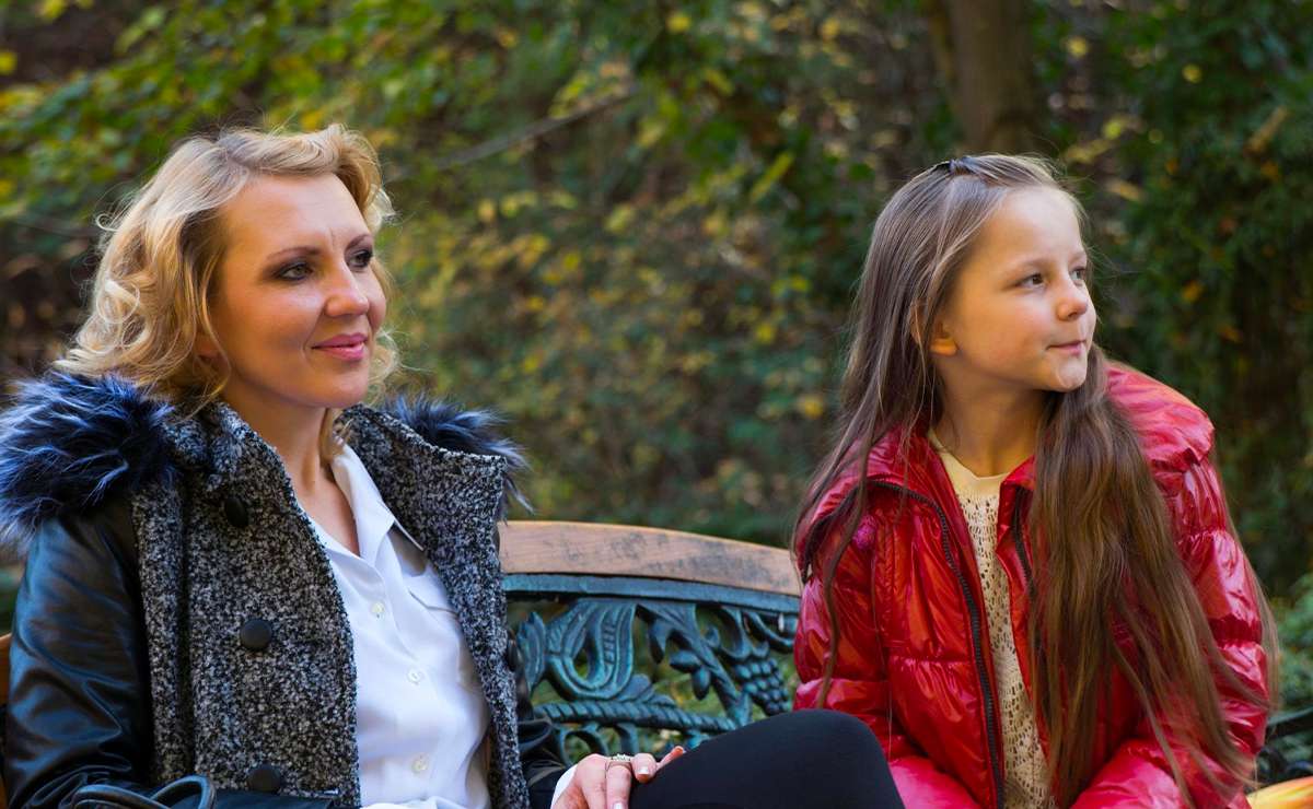 Donna bionda e bambina con giacca rossa sedute su una panchina in ferro battuto, immerse in un ambiente autunnale nel verde
