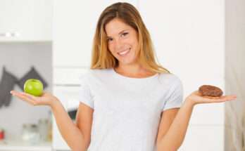 Donna sorridente che tiene in mano una mela verde e una ciambella al cioccolato, simbolo della scelta tra alimenti salutari e confort food nei momenti di calo dell’umore