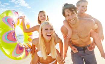 Famiglia sorridente in spiaggia con due bambini, salvagente colorato e mare sullo sfondo: vacanze estive in Campania a misura di bambini.