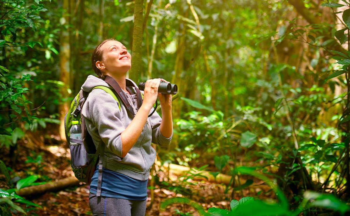 Donna con zaino e binocolo che osserva gli uccelli in una foresta tropicale, durante un’escursione di birdwatching immersa nella natura.