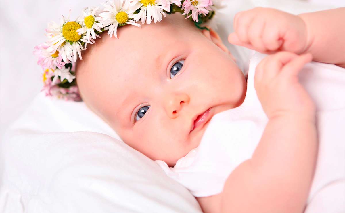 Bambina con occhi azzurri e coroncina di margherite rosa e bianche, sdraiata su un lenzuolo bianco