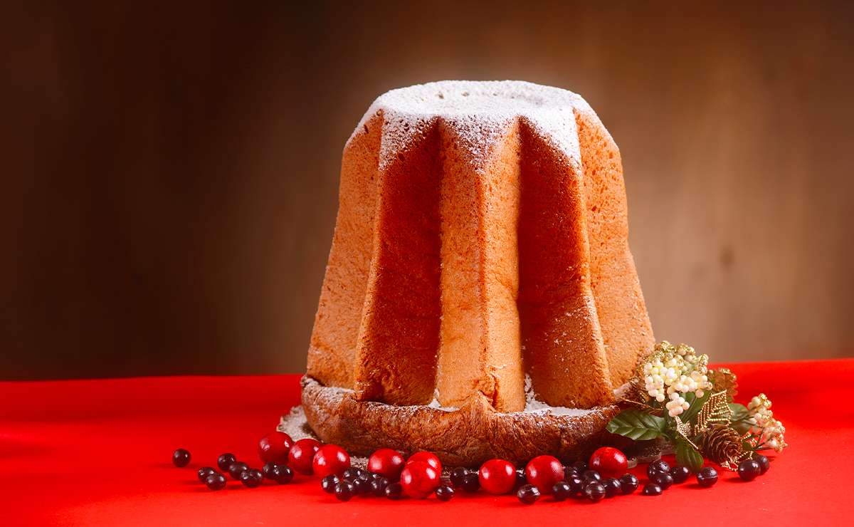 Pandoro classico con zucchero a velo su tovaglia rossa, decorazioni natalizie con bacche rosse, pigne e fiori sullo sfondo.