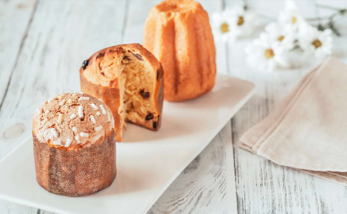 Mini panettone, panettone classico e pandoro serviti su piatto bianco, con tovagliolo beige e fiori bianchi decorativi su sfondo in legno chiaro.