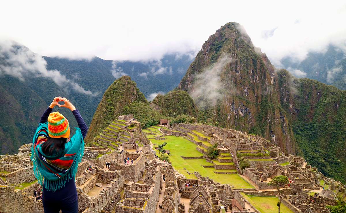 Turista con poncho colorato e berretto tipico che forma un cuore con le mani davanti alle rovine di Machu Picchu incorniciate dalle montagne andine nebbiose.