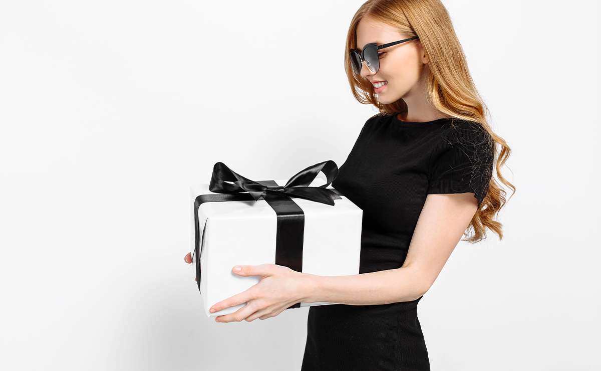 Donna con abito nero che sorride mentre tiene in mano un pacco regalo bianco con fiocco nero, perfetto per il Black Friday