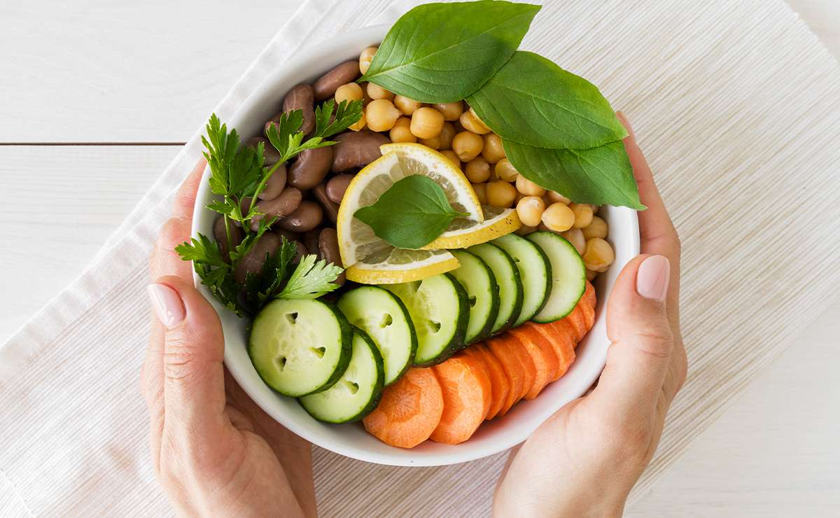 Ciotola vegana con fagioli, ceci, cetrioli, carote, fette di limone e foglie di basilico, tenuta tra le mani su tovaglietta chiara.