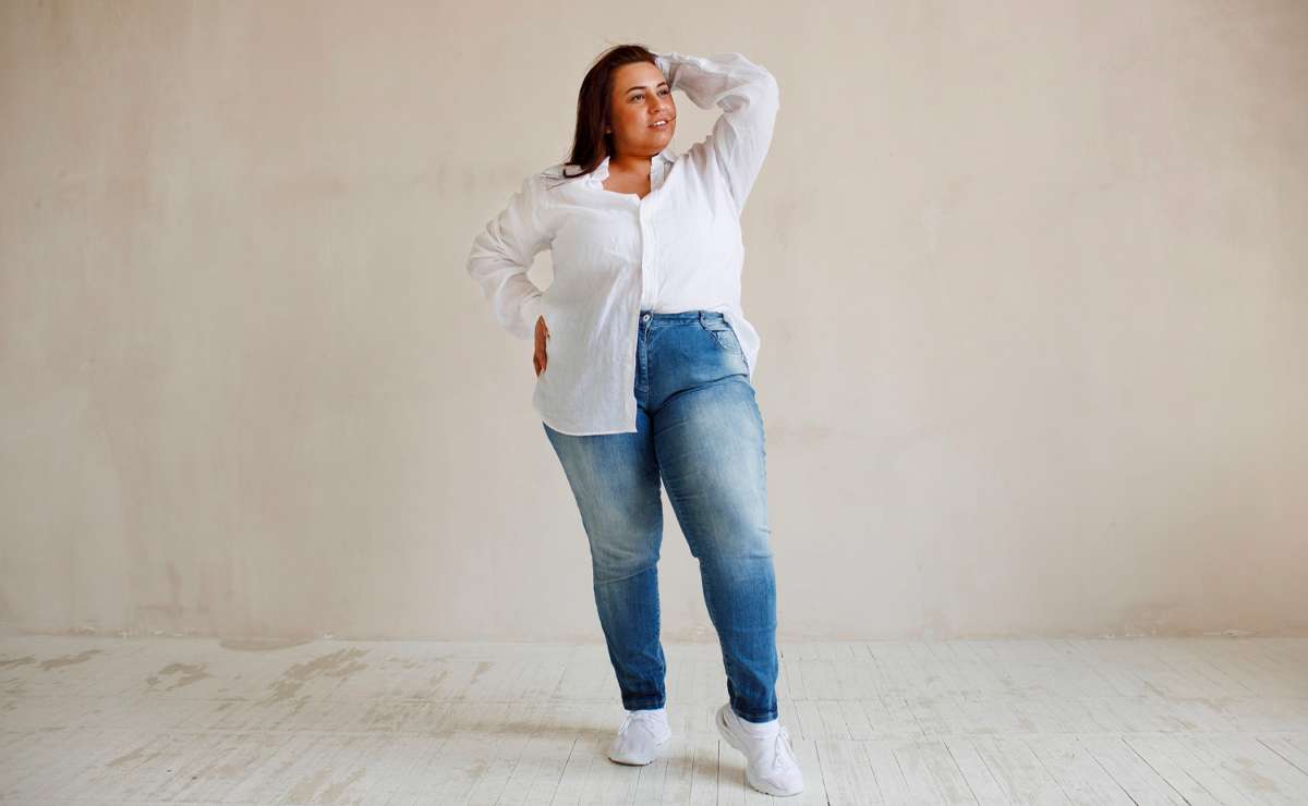 Donna curvy posa con sicurezza indossando jeans a vita alta e camicia bianca, in un ambiente minimalista
