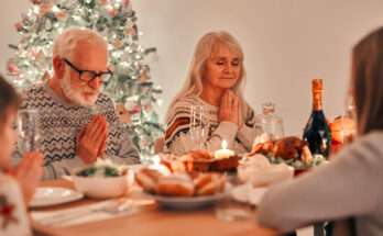 Coppia di anziani cristiani che pregano a tavola durante la cena di Natale, con albero addobbato e candele accese, simbolo della celebrazione religiosa.