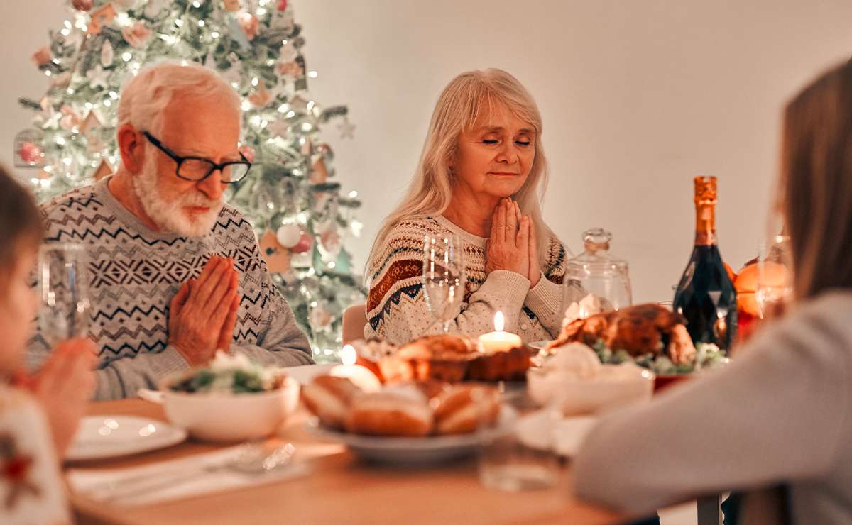 Coppia di anziani cristiani che pregano a tavola durante la cena di Natale, con albero addobbato e candele accese, simbolo della celebrazione religiosa.