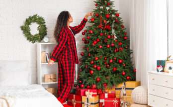 Donna in pigiama rosso e nero che decora l’albero di Natale con palline rosse e luci, circondata da regali colorati sotto l’albero.