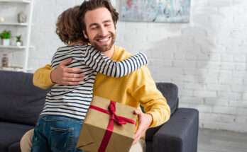 Papà che riceve un regalo e abbraccia il figlio sorridendo sul divano, momento felice e affettuoso per un’idea regalo originale e personalizzata.