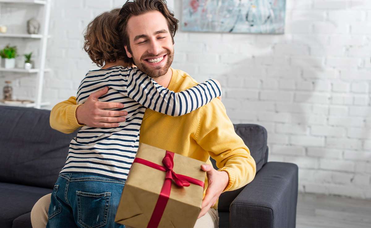 Papà che riceve un regalo e abbraccia il figlio sorridendo sul divano, momento felice e affettuoso per un’idea regalo originale e personalizzata.