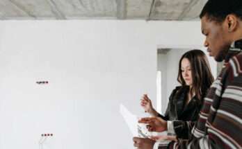 Due persone controllano dei documenti all’interno di una casa in ristrutturazione, con pareti grezze e impianti elettrici visibili.