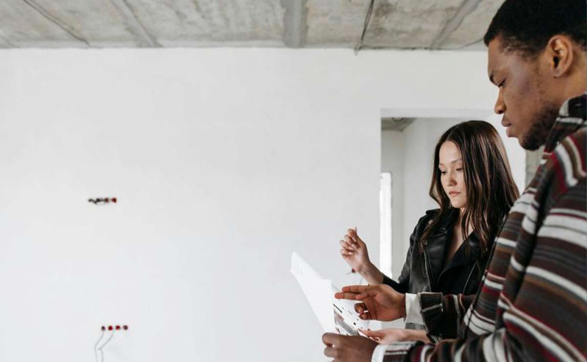 Due persone controllano dei documenti all’interno di una casa in ristrutturazione, con pareti grezze e impianti elettrici visibili.