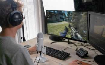 Ragazzo con cuffie e microfono che gioca a un videogioco sparatutto al computer, con doppio monitor e tastiera su una scrivania.