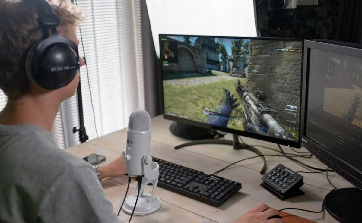 Ragazzo con cuffie e microfono che gioca a un videogioco sparatutto al computer, con doppio monitor e tastiera su una scrivania.