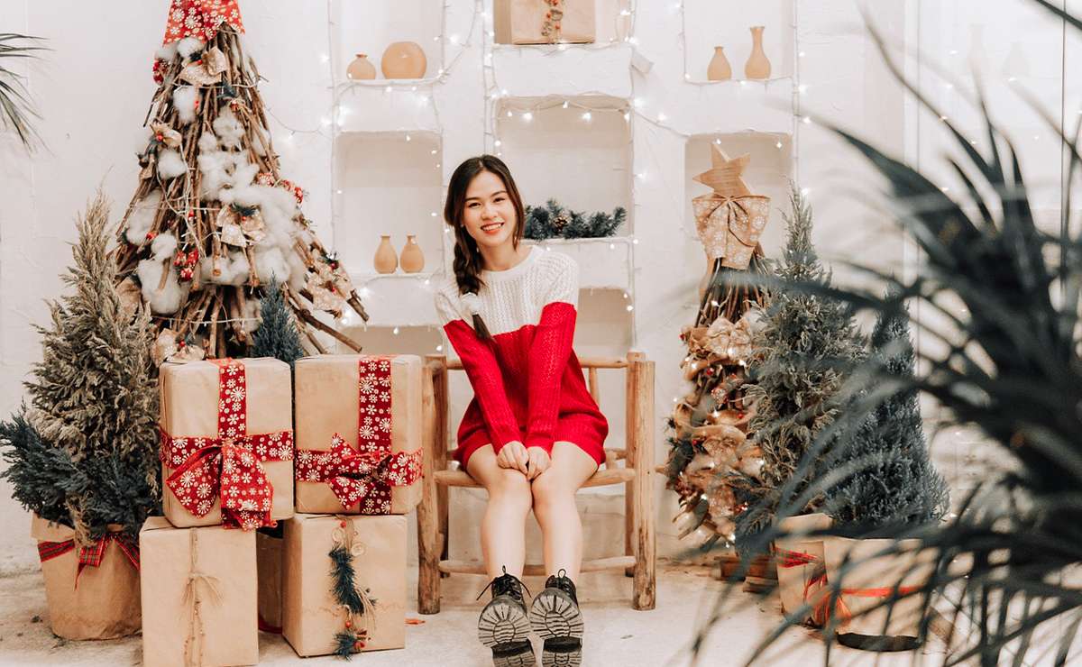 Donna sorridente in abito rosso natalizio seduta tra alberi decorati e pacchi regalo