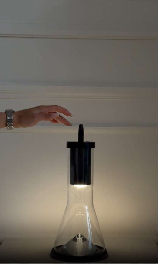 Mano che si avvicina a una lampada di design con base in vetro e luce calda accesa, in un ambiente moderno e minimalista.