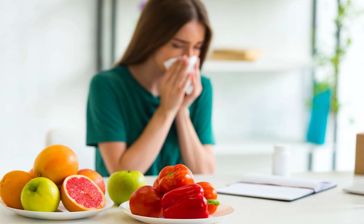 Donna raffreddata seduta al tavolo che si soffia il naso, con un piatto di agrumi e un piatto di peperoni ricchi di vitamina C in primo piano