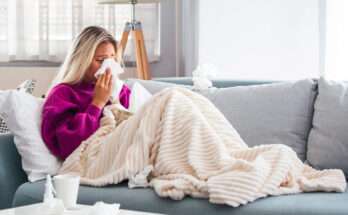 Donna sul divano con il raffreddore che si soffia il naso, avvolta in una coperta chiara con fazzoletti usati accanto.