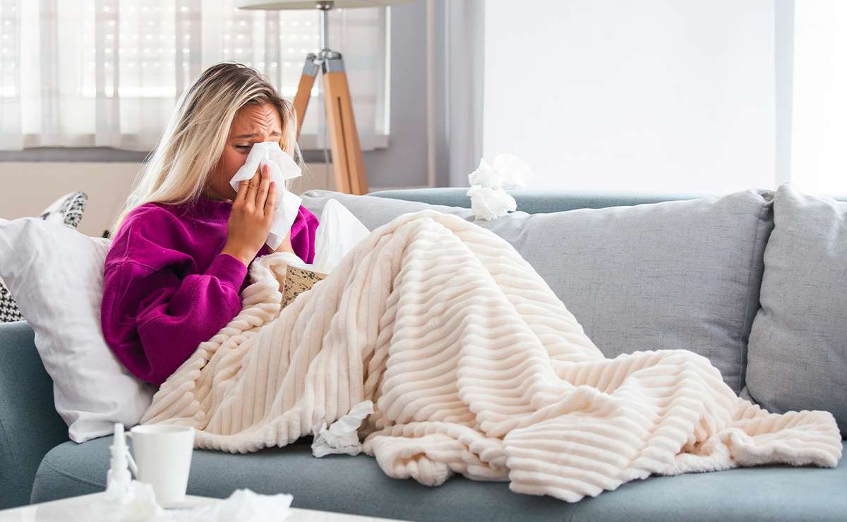 Donna sul divano con il raffreddore che si soffia il naso, avvolta in una coperta chiara con fazzoletti usati accanto.
