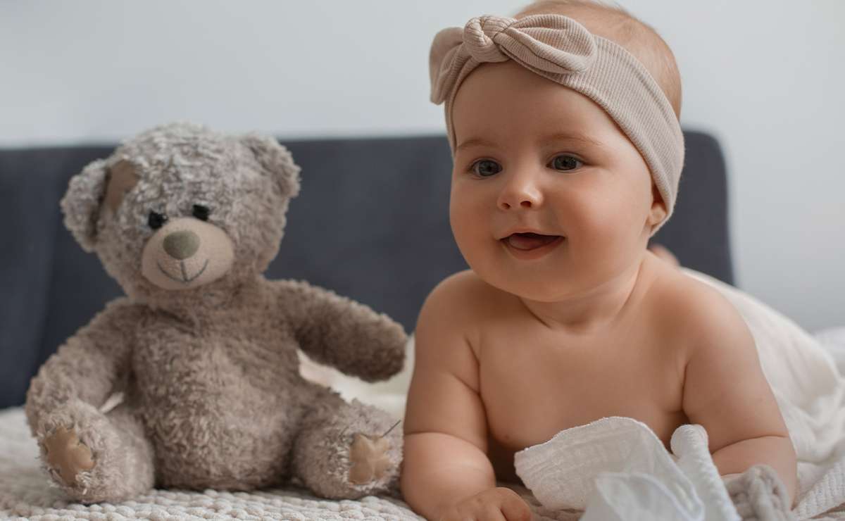 Bambina con fascia beige in testa che sorride sdraiata accanto a un peluche