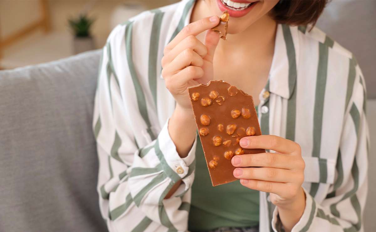 Donna che mangia una tavoletta di cioccolato al latte con nocciole, seduta sul divano con camicia a righe verdi e bianche.